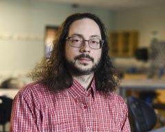 Professor Jonathan Gaffney, with glasses, beard and hair to his shoulders, dressed in a red shirt, stands in his classroom.