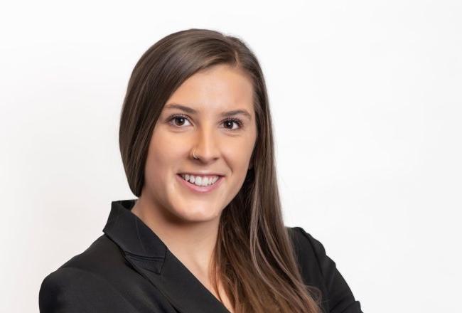 Madison Judge stands with arms folded in a black blazer, smiling at the camera against a white background.